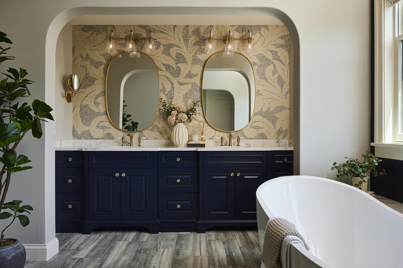 This primary bathroom transformation focused on refreshing a dated space while improving functionality and preserving quality craftsmanship. Existing cabinetry was updated with paint, while new tile, a custom half tub deck, and a freestanding tub brought a lighter, more modern aesthetic. The redesigned space highlights the barrel-vaulted ceiling with statement wallpaper and is finished with brass and crystal accents that add warmth, elegance, and personality.