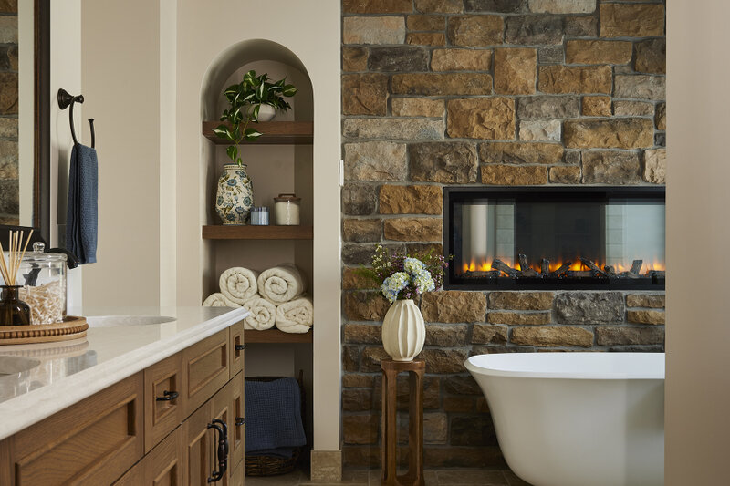 This primary bathroom remodel in Blaine, MN, combines warm wood cabinetry, a stone fireplace, and a freestanding soaking tub for a cozy, spa-inspired retreat. A zero-entry walk-in shower adds comfort and accessibility to the refreshed space.